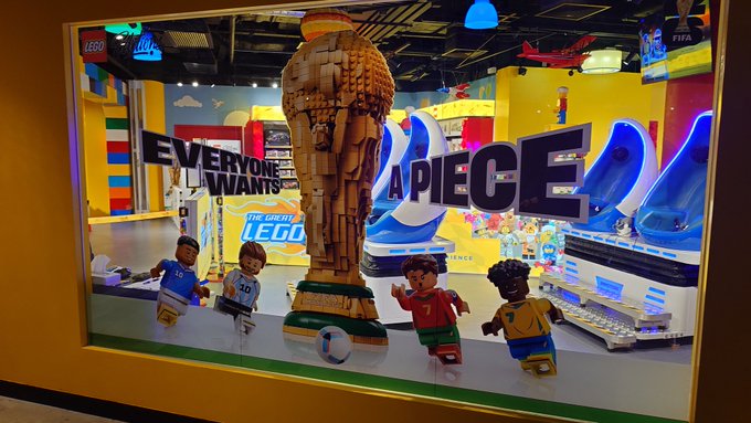 A LEGO display window features a large golden LEGO World Cup trophy with the text "EVERYONE WANTS A PIECE" alongside LEGO minifigures of soccer players, connecting to the poster's theme of visiting a World Cup LEGO exhibition in Hong Kong on a 32°C day.