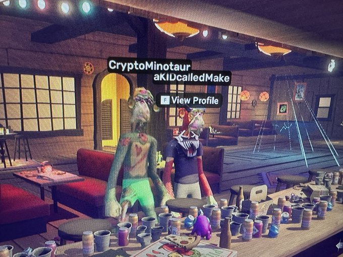 A video game screenshot showing two characters, 'CryptoMinotaur' and 'aKIDcalledMake', standing behind a counter filled with mugs in a cozy room.