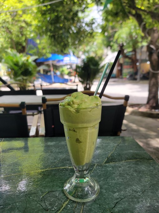 A green avocado or matcha blended drink in a tall sundae glass with two straws, sitting on a green marble table at an outdoor cafe.