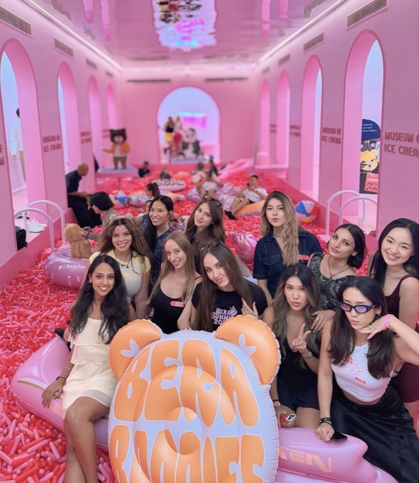 A group photo of several young women posing together in a pink-themed Museum of Ice Cream, sitting in a ball pit with a large "BEAR BUBBLES" inflatable prop in the foreground.