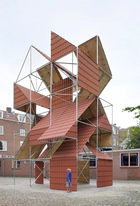 A tall architectural pavilion structure constructed from red-tiled roof panels arranged in angular geometric formations on a metal frame stands in a courtyard.