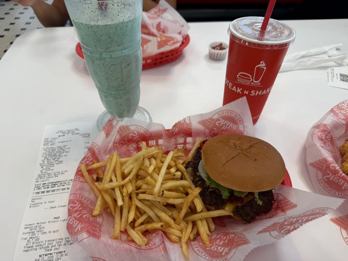A meal at a diner table features a burger, fries in a red-lined basket, a tall blue milkshake, and a red 'STEAK N SHAKE' cup with a straw. A receipt is visible on the table next to the food.