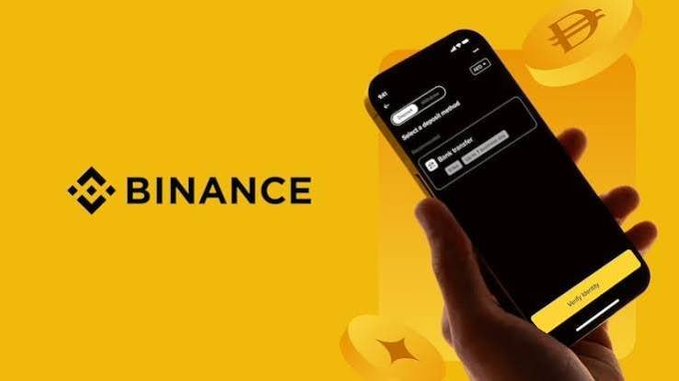 A hand holds a smartphone displaying the Binance app with a "Borrow" option and fields for "Select a crypto asset" and "Borrow transfer," set against a vibrant yellow background featuring the Binance logo and cryptocurrency coin graphics—illustrating the platform's crypto-backed borrowing feature highlighted by the post.