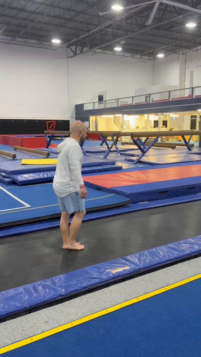 The image features a man standing barefoot on a blue mat inside an indoor gym, wearing shorts and a white shirt. The man appears to be looking at his phone or possibly observing something else in the room. There are several exercise machines around him, including a bench press and a pull-up bar. A yellow line is visible on the floor near the man's feet, which could indicate a designated area for specific exercises or activities within the gym.