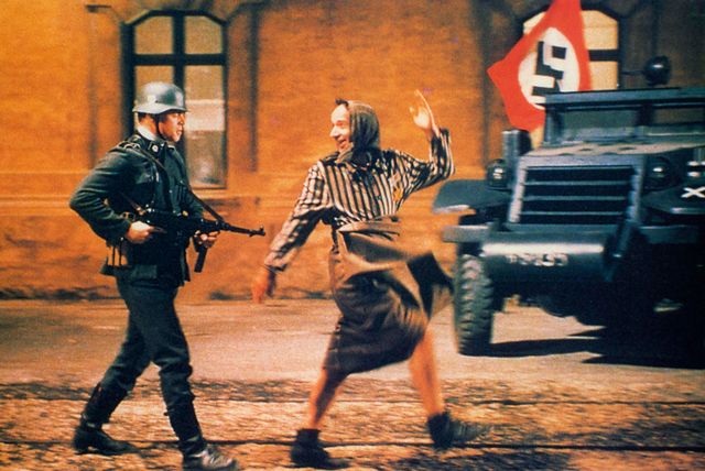 A cinematic still showing a Nazi soldier with a rifle facing a dancing woman in striped clothing, with an armored vehicle and swastika flag visible in the background.