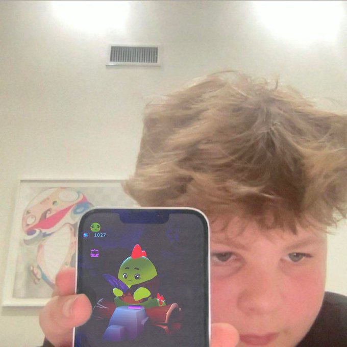 The image features a young man holding up his cell phone with a cartoon character displayed on its screen. The cartoon character appears to be riding a car, which adds an element of fun and entertainment to the scene. The boy seems to be taking a selfie or sharing something interesting he found on his phone with others.