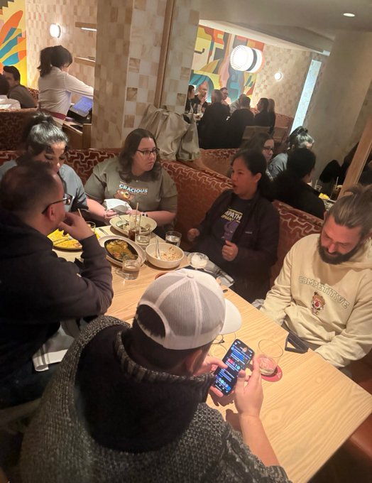 A group of people sit around a wooden table in a restaurant booth, with one person in a gray cap holding a phone displaying an app.