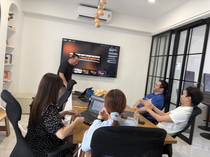 A group of five people are gathered in a meeting room with laptops, collaborating while viewing a presentation on a wall-mounted TV displaying text including 'DON'T SEND ME MONEY' and 'IMPACT WITH US'.