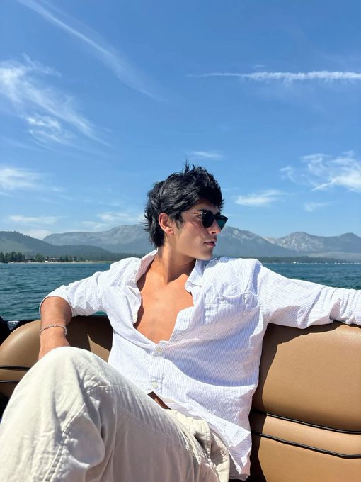 A young man wearing an open white striped shirt and sunglasses sits on a boat with mountains and water in the background.