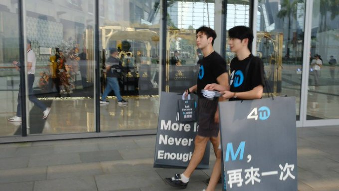 Two young men walk past a glass building carrying signs referencing "4π" (likely related to the user @4MBossChen mentioned in the post), with one sign reading "More Is Never Enough" and another displaying "M, 再来一次" ("M, one more time"), visually playing on the 4MBossChen meme by highlighting the bold, attention-grabbing promotion surrounding the exchange's launch. The casual street setting reinforces the post's skepticism about the hype, contrasting flashy signage with the practical concerns over trade execution, asset security, and accountability.