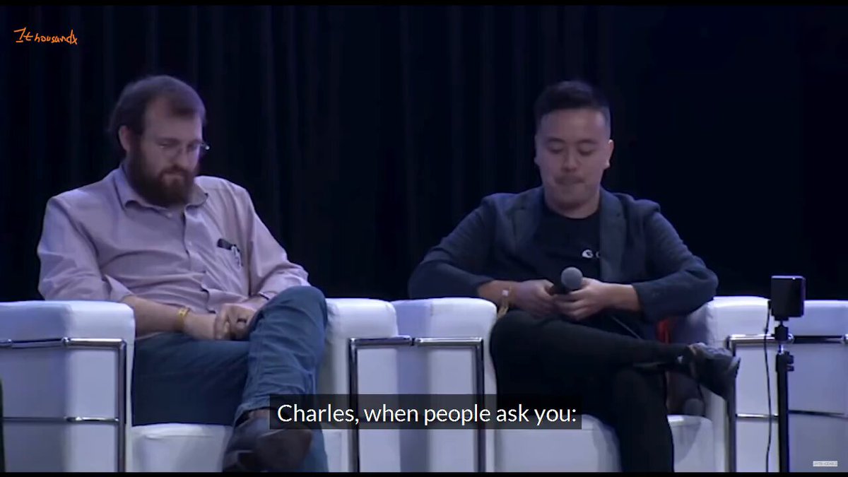 The image shows two men sitting next to each other on white chairs with their legs crossed. One man has a beard and glasses, while the other has short hair and is wearing a black shirt. They are both looking at something off-camera, possibly engaged in a conversation or presentation. The text "Charles, when people ask you" can be seen near them, suggesting that they might be discussing a topic related to Charles Hoskinson's work or interests.