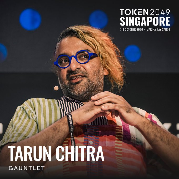 A speaker wearing blue glasses and a striped shirt gestures with clasped hands at a TOKEN2049 Singapore panel, with the event graphic identifying him as "TARUN CHITRA, GAUNTLET" and dated "7-8 OCTOBER 2026 · MARINA BAY SANDS," promoting his return appearance at the conference where he leads the DeFi risk management firm.