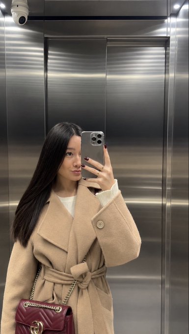 A mirror selfie of a woman with long black hair wearing a camel-colored coat over a white top, carrying a burgundy quilted Chanel handbag while standing in an elevator.