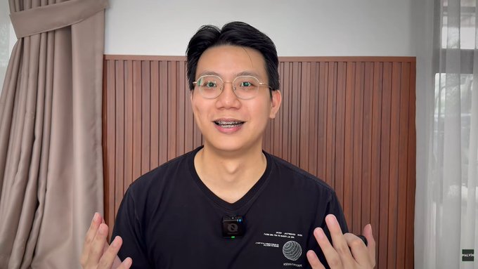 A man wearing glasses and a black t-shirt with a microphone clipped to it stands against a wooden panel wall with curtains, speaking with his hands raised.