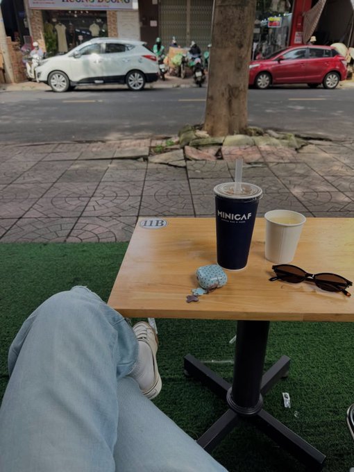 First-person view at an outdoor café table with a MINICAFF drink, a paper cup, sunglasses, and a small toy car, with a street and parked cars visible in the background.