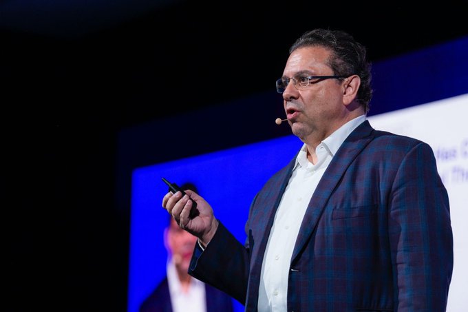 The image shows a man wearing glasses standing on stage, holding a microphone and speaking into it. The background features a large screen displaying information about a conference or event. The caption of the image reads "Focus on what is in the image and how it relates to the post."