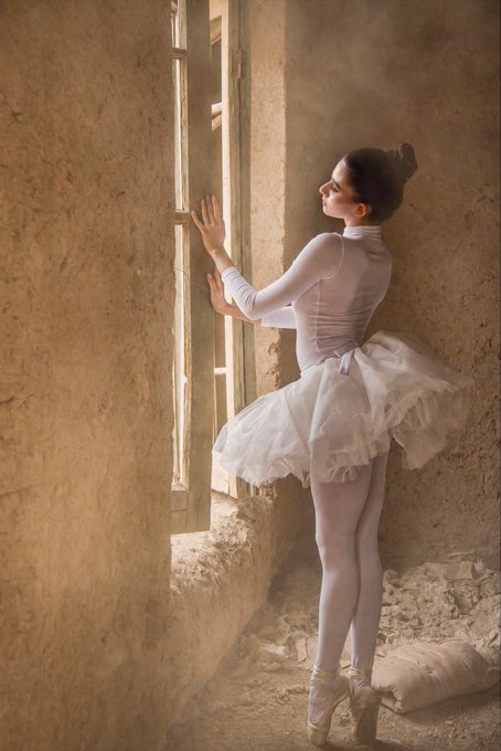 A ballerina in a white tutu and pointe shoes standing en pointe in a dilapidated room, reaching toward a sunlit window with textured stone walls.