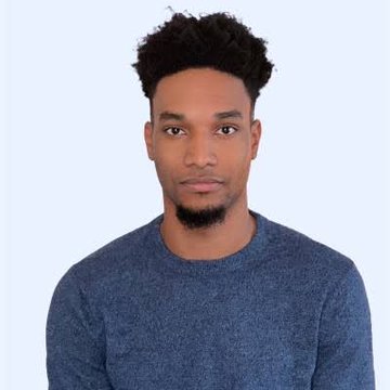 A young man with short curly hair and a small goatee wearing a blue crewneck sweater against a light blue background.