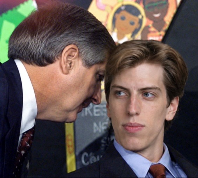 The caption should focus on what is visible in the image without making assumptions about identity or context.

An older man in a dark suit leans in close to whisper into the ear of a younger man with brown hair, who wears a dark suit and tie and gazes forward with a blank, impassive expression. Colorful artwork is visible in the background, but no clear text or identifiers can be read.