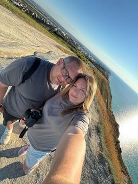 A couple takes a selfie on a coastal cliff, with a sandy beach, ocean, and distant town visible behind them under a clear sky.