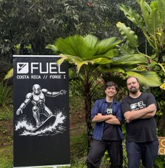 Two men stand proudly with arms crossed beside a black banner featuring a muscular astronaut illustration and the text "FUEL COSTA RICA // FUEL" at what appears to be an outdoor event in Costa Rica, with lush tropical foliage in the background. The image relates to the post about FUEL's continued presence and growth, showcasing co-founders including Nick Dodson, Rishabh Keshan, and voxelot who are "still here to build" amid organizational changes.