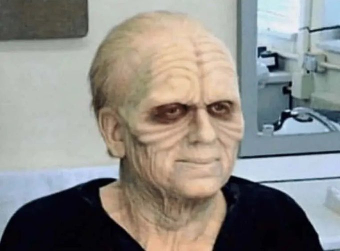 An older man sits in what appears to be a bathroom or salon, wearing heavy zombie/undead makeup with pale greenish-gray skin, sunken dark eyes, and weathered facial features. The image humorously illustrates the post's sentiment about feeling like a decaying corpse after a rough morning hangover, with no visible text or numbers in the image itself.