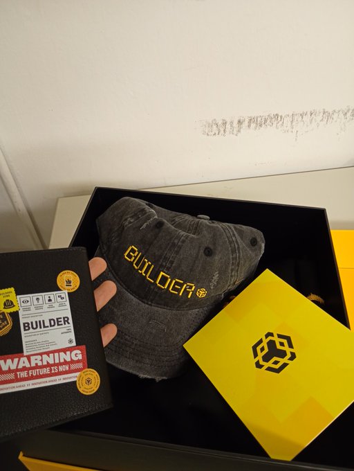 The image features a black hat with yellow text that reads "builder", placed inside of a box, along with some other items such as a book and a warning label. The scene appears to be set against a white wall or background, which highlights the hat and its contents.