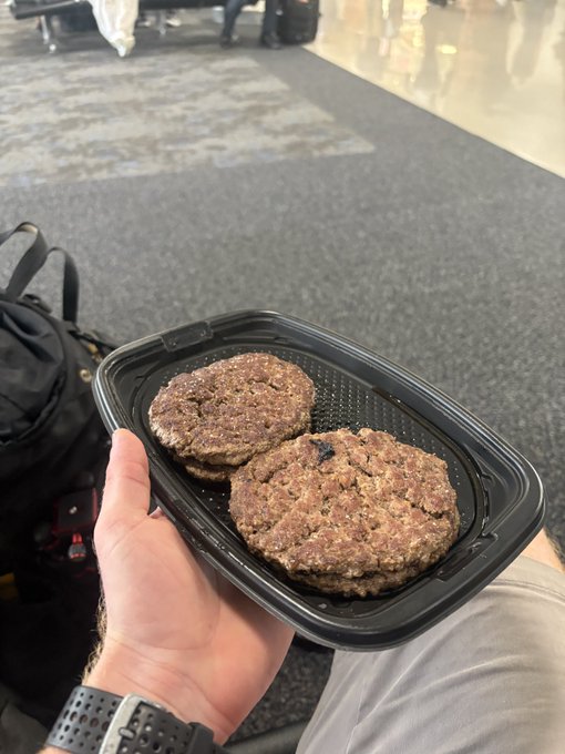 A traveler holds a black McDonald's clamshell container with two quarter-pound beef patties, likely at an airport based on the luggage and tiled flooring in the background. The post references four patties total for $9, suggesting two containers were purchased—a tip from economist Saifedean Ammous about cost-effective protein while traveling.