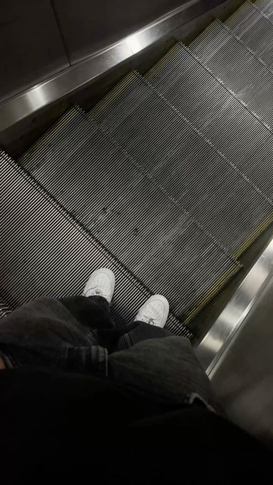 A first-person perspective shot looking down at a person's feet wearing white sneakers on a descending metal escalator, with the edge of denim jeans visible.