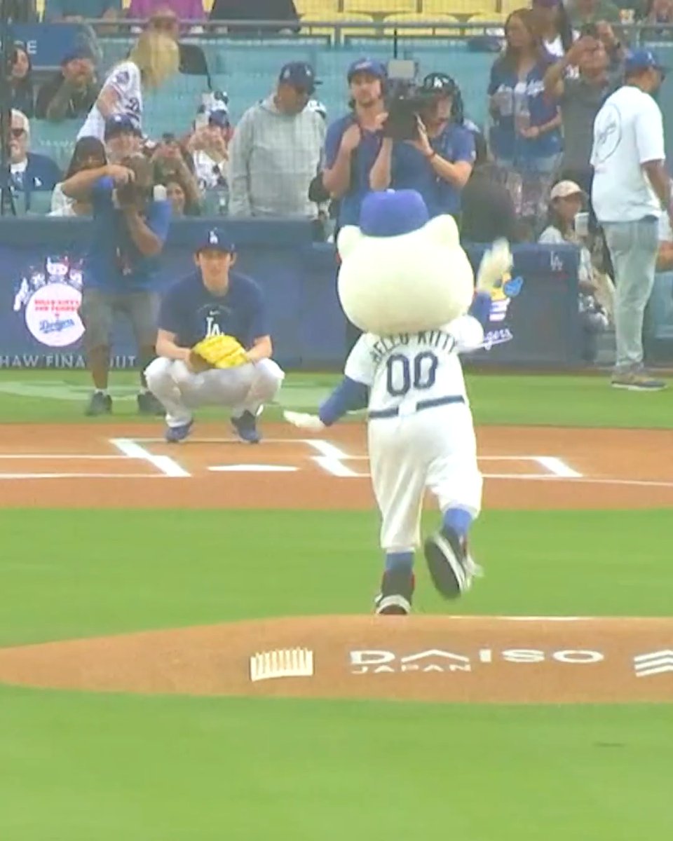 The Hello Kitty mascot wearing a Dodgers baseball uniform with 'HELLO KITTY' and '00' on the back, running onto a baseball field at Dodger Stadium, with players and photographers in the background.