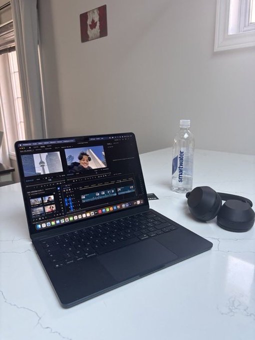 The image shows a laptop computer sitting on top of a white table with a water bottle next to it, as well as headphones nearby. The laptop screen displays a video editing software interface, suggesting that someone might be working on a project or creating content for social media. The presence of the water bottle and headphones indicates that this person is likely spending an extended period at their desk, possibly working from home or engaging in creative activities like video editing.
