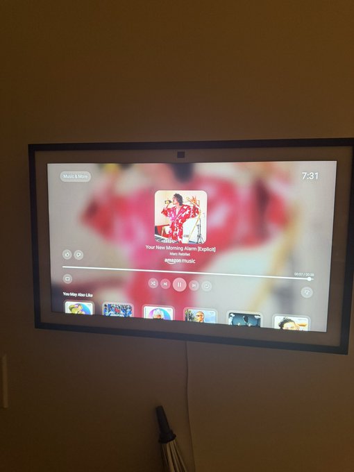 A wall-mounted smart display showing the Apple Music interface, currently playing an album titled 'Your New Morning Alarm (Explicit)' from an Amazon Music playlist at 7:31.