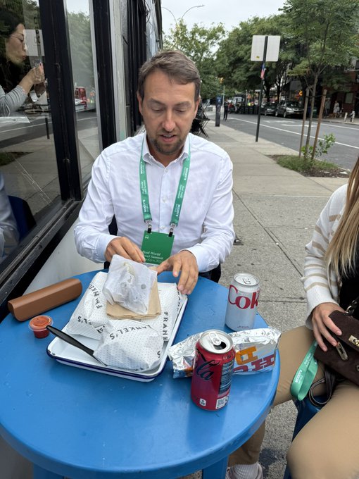 A man in a white shirt and lanyard unwraps a sandwich on a blue outdoor table alongside a Coca-Cola can and a red beverage, with a city street visible behind him. The scene captures a casual moment during the RWA Summit referenced in the post, reflecting the on-the-ground energy of the institutional crypto event.