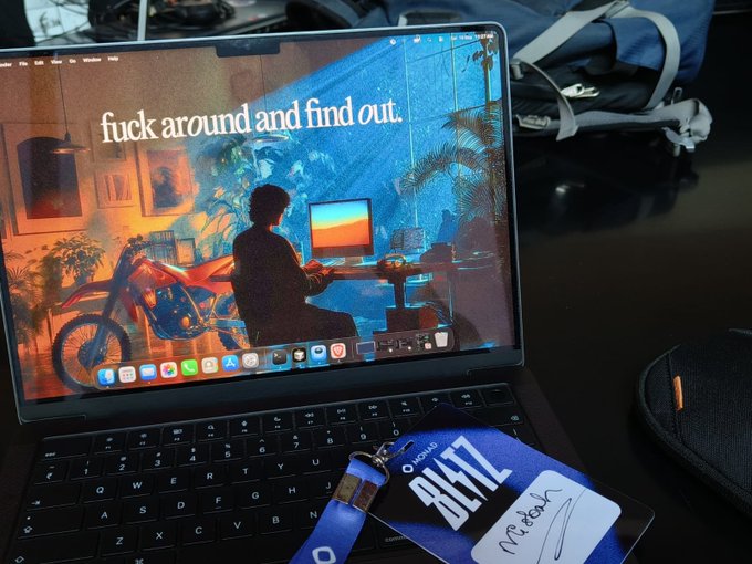 A photograph of an open laptop displaying a desktop wallpaper with the text 'fuck around and find out,' alongside an Apple lanyard badge with the name 'Rietzl' and a stylus.