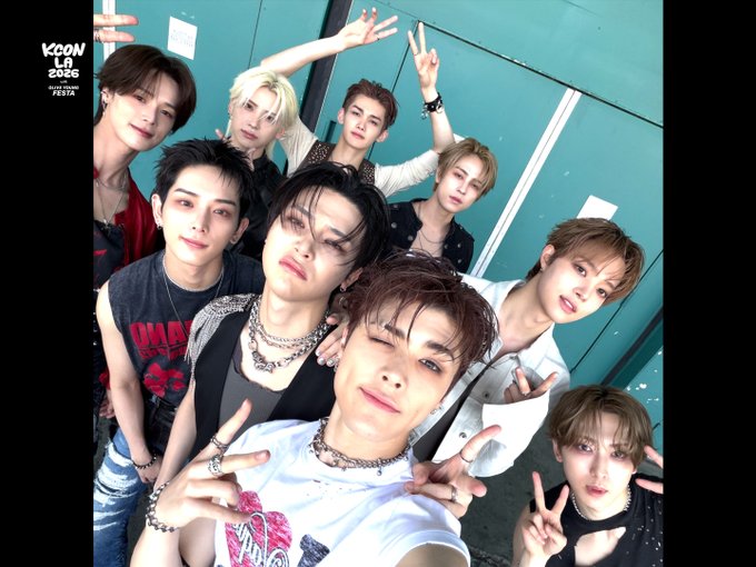 The image shows members of the Japanese group &TEAM posing for a greenroom selfie at KCON LA 2026, with the "KCON LA 2026" logo visible in the top left corner. Nine members are gathered closely together, smiling and making peace signs, capturing an energetic behind-the-scenes moment from the group's Day 1 appearance on August 14.
