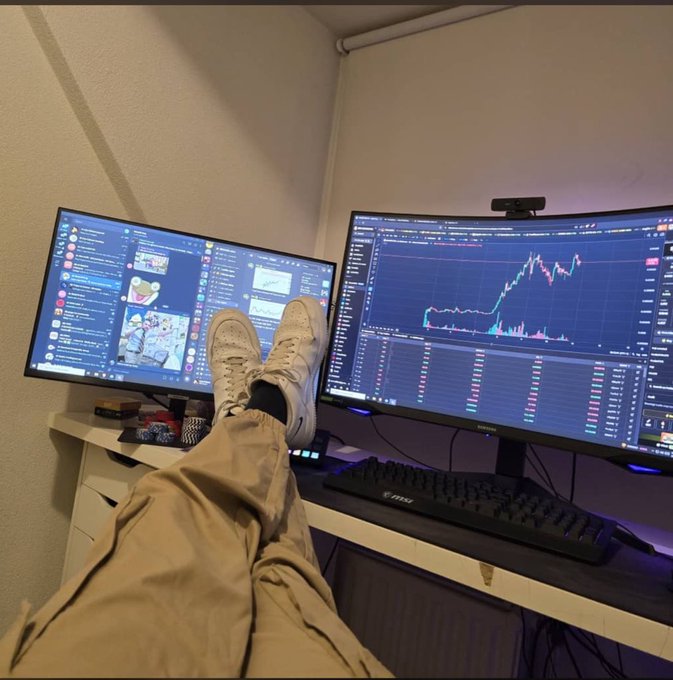 A first-person view shows legs in khaki pants and white sneakers propped on a desk with two monitors—one displaying chat and another showing a crypto trading chart.