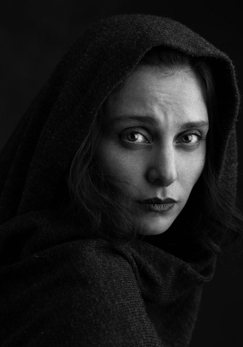 Black and white portrait of a woman with an intense gaze, wearing a dark hooded cloak.