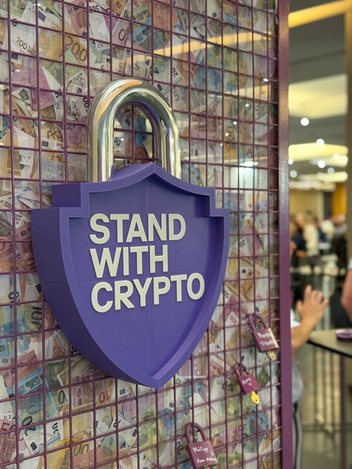 A purple padlock-shaped sign reading 'STAND WITH CRYPTO' mounted on a wall covered in various international banknotes displayed in a wire grid.