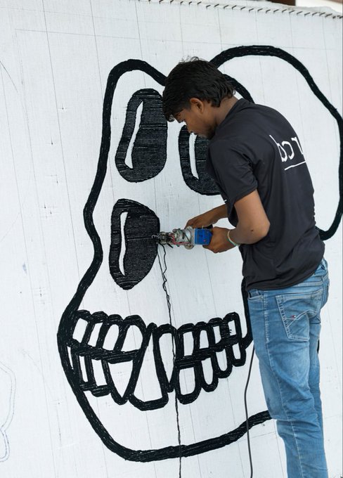 A young man in a black t-shirt and blue jeans using a tool to carve or draw a large black skull design onto a white wall.