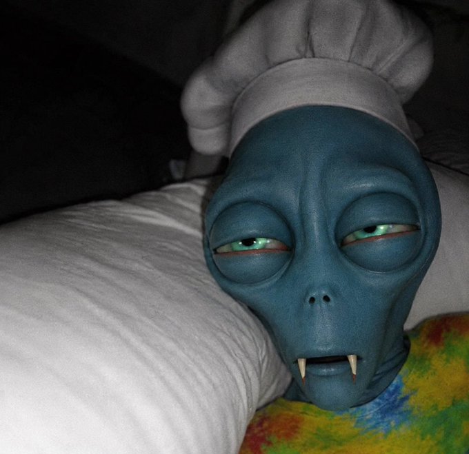 A blue, alien-like mask with fangs and a white chef's hat resting against a pillow in dim lighting.