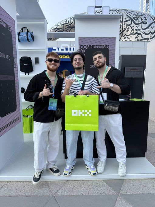 Three men pose at an OKX booth with an 'OKX' branded green bag, with backdrop signage reading 'CONNECTING' and 'Connecting You' visible behind them.
