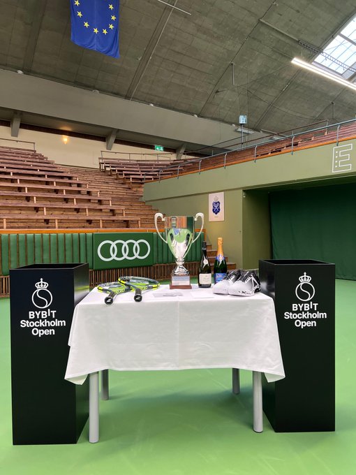 A table set up on a tennis court displays the Bybit Stockholm Open trophies, racquets, and champagne bottles, flanked by two black podiums branded with "BYBIT Stockholm Open" logos. The display, positioned beneath an EU flag and a Champions Trophy banner, captures the pre-tournament atmosphere at Kungliga Tennishallen as described in the post.