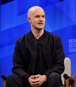 A bald man in a black jacket sits on a stage with a blue background that partially reads 'NEW ESTATE SUMMIT'.