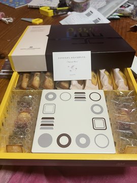 The image shows a box of chocolates sitting on top of a table, with a note attached to it that reads "あけるおめそうございます スーパー". The box contains various types of chocolate bars and is placed next to a yellow tray.