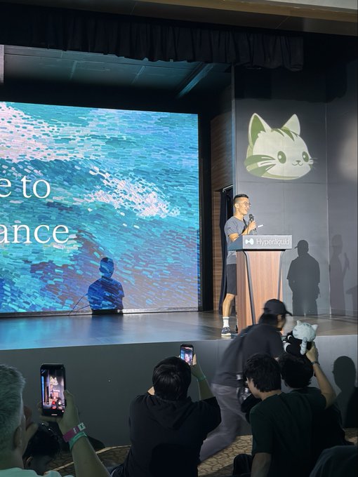 A man speaks at a podium branded with "Hyperliquid" at what appears to be a conference or product launch event, with a large screen displaying ocean imagery and partial text reading "e to" and "ance" visible behind him. The Hyperliquid logo (a cat illustration) is projected on the wall, and audience members are recording the presentation on their phones, aligning with the post's reference to a "father" figure speaking about Hyperliquid.