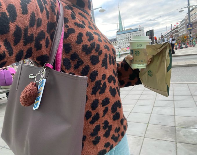 A person wearing a leopard print sweater carries a gray handbag with a fuzzy brown charm and a Starbucks keychain, while holding a green Starbucks drink and a paper bag in a European city setting.