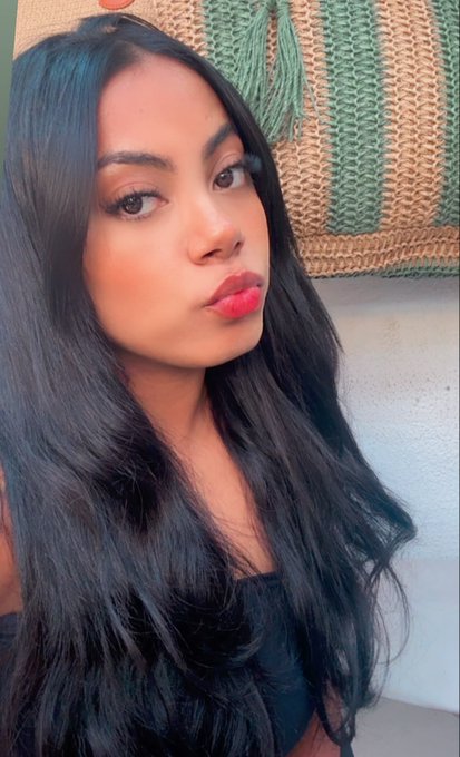 A close-up selfie of a woman with long dark hair, red lipstick, and makeup, posing in front of a woven wall hanging with green tassels.