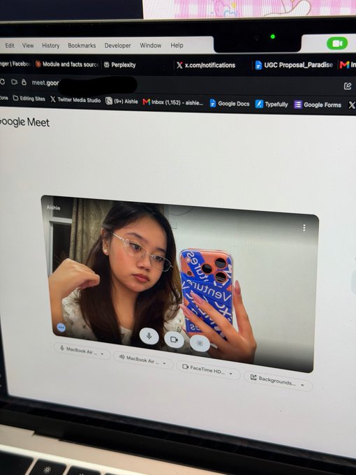 A young woman with glasses takes a mirror selfie on a MacBook Air during a Google Meet call, her blue phone case featuring "Eventbrite" branding visible as she rests her chin on her hand with a tired expression. The image captures a relatable "gm" (good morning) moment of delayed morning motivation, showing her logging on virtually but not quite ready to start the day with full discipline.