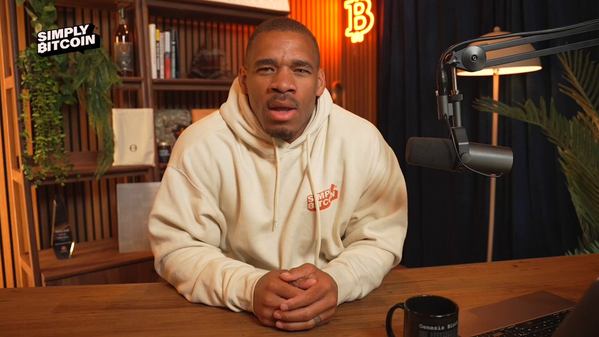 The image shows a man sitting at a desk with a microphone in front of him. He appears to be wearing a white hoodie and has his hands folded in front of him, suggesting that he might be participating in an interview or speaking event. The background features a bookshelf filled with various items such as vases and potted plants, giving the impression of a well-decorated room. There is also a laptop on the desk, which could be used for recording or streaming the event.