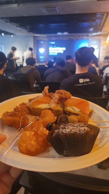 A plate of assorted desserts including a chocolate muffin, orange slices, and pastries is held up in the foreground at a tech event, with a blurred audience watching a presentation on a screen in the background.